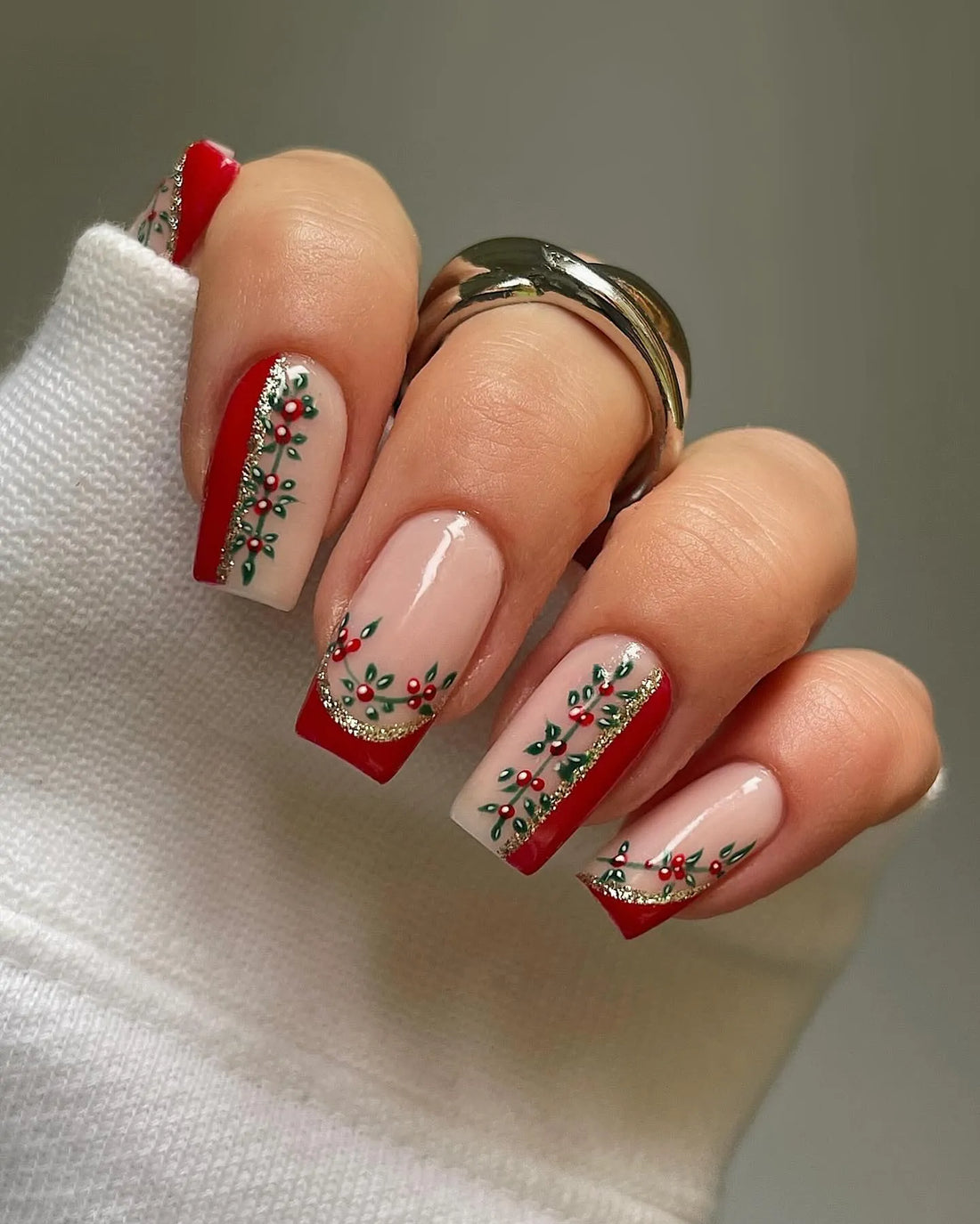 Hand with red and nude holiday press-on nails, holly vine and glitter Christmas nail art.