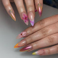Hand with iridescent pink chrome press-on nails, 3D floral accents, and sunset ombre tips