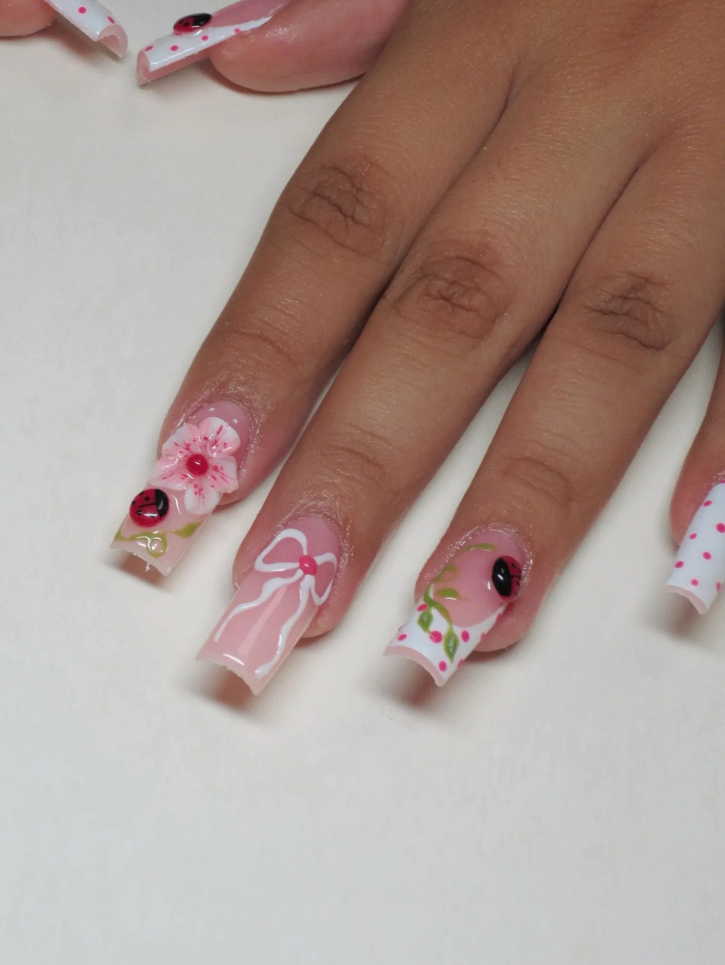 Hand with handmade pink press-on nails featuring 3D floral, ladybug, bow, and polka dot designs.