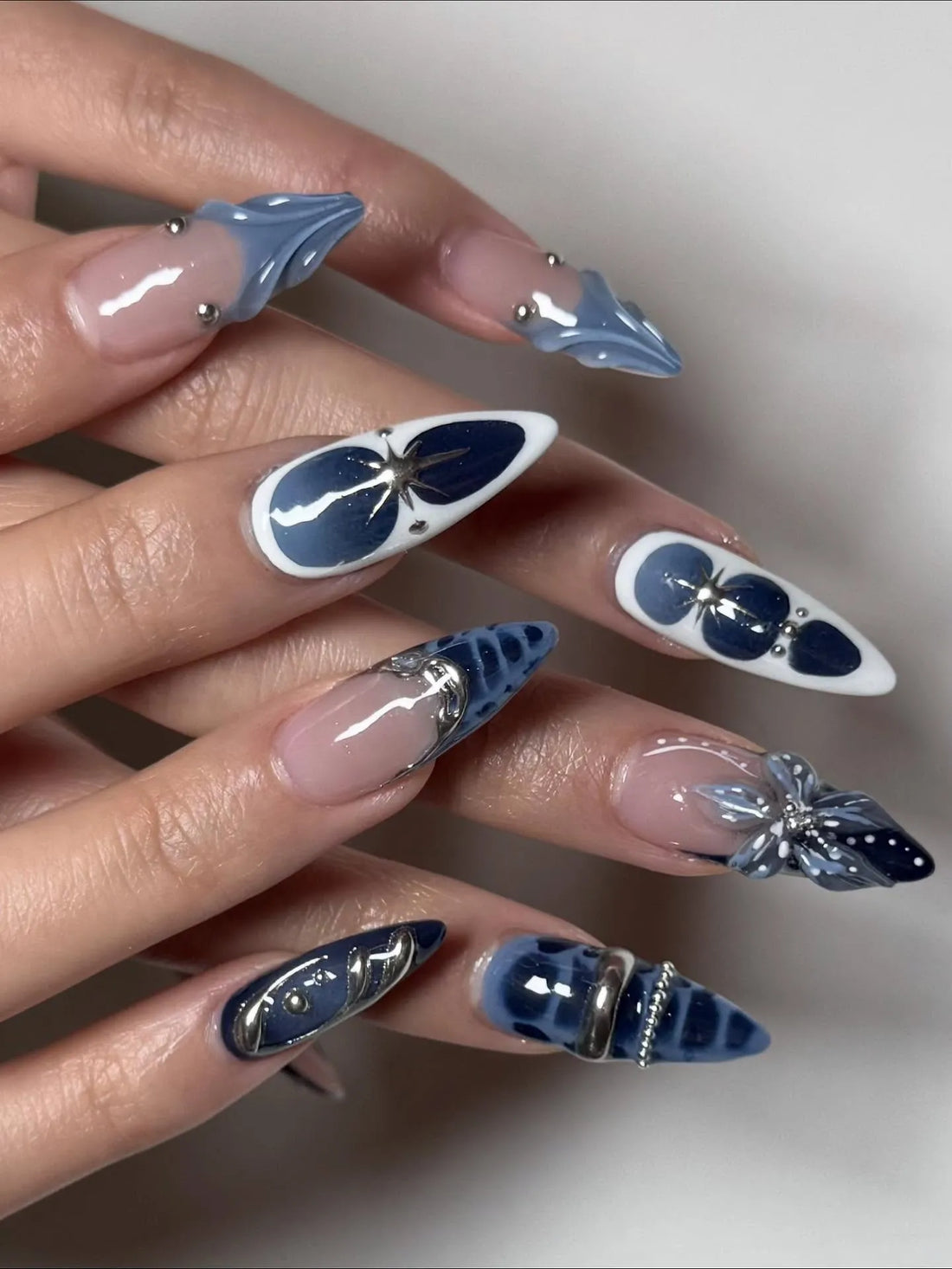Hand with long stiletto press-on nails in midnight blue, silver chrome, 3D floral and celestial designs.