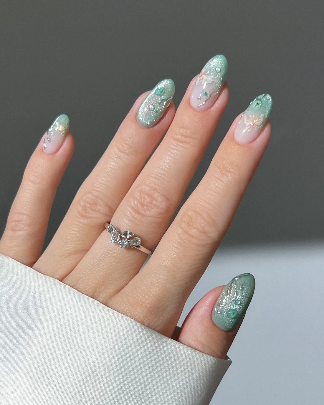 Hand with mint green ombre press-on nails featuring 3D butterfly, floral, and glitter accents.