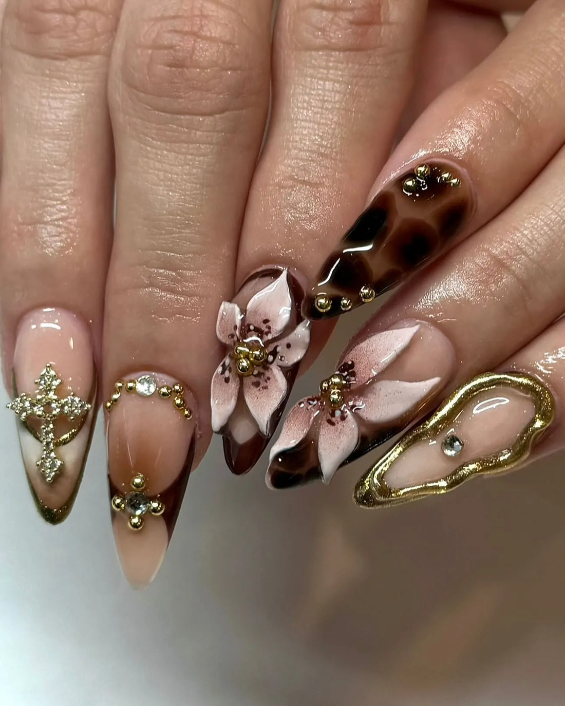 Hand with handmade press-on nails featuring mocha leopard print, gold swirls, 3D floral and rhinestone accents.