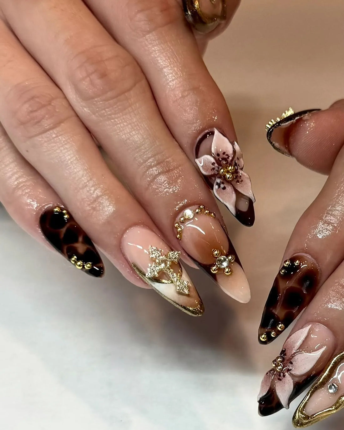 Hand with handmade press-on nails featuring mocha leopard print, gold swirls, 3D floral and rhinestone accents.