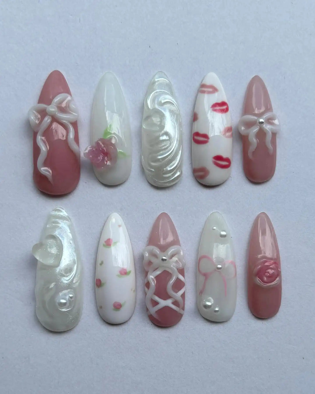 Handmade press-on nails with pink bows, roses, lip prints, lace and heart designs on white background.