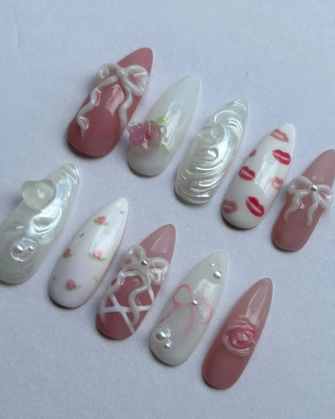 Handmade press-on nails with pink bows, roses, lip prints, lace and heart designs on white background.