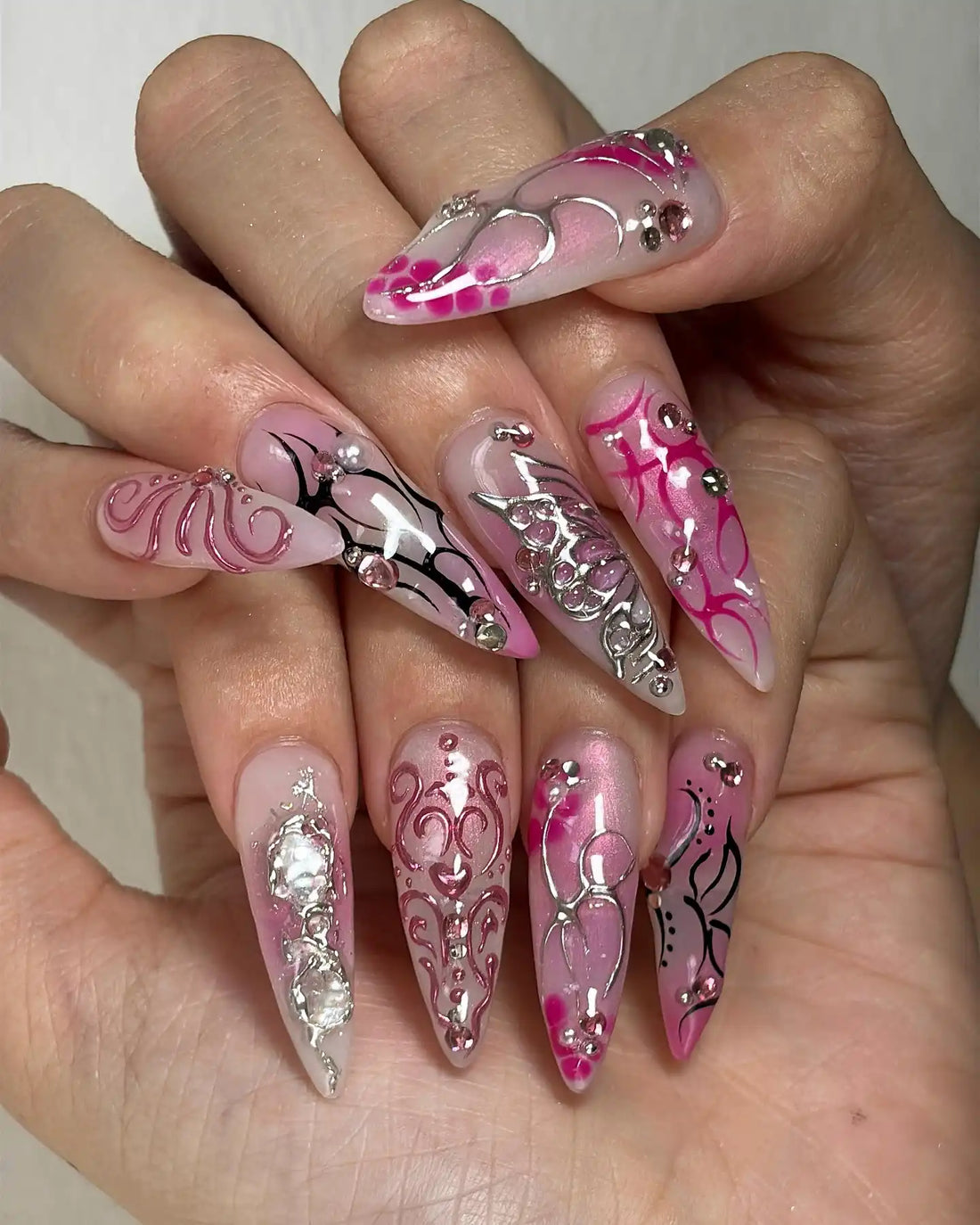 Hand with long stiletto press-on nails featuring pink, chrome, and silver rhinestone designs.
