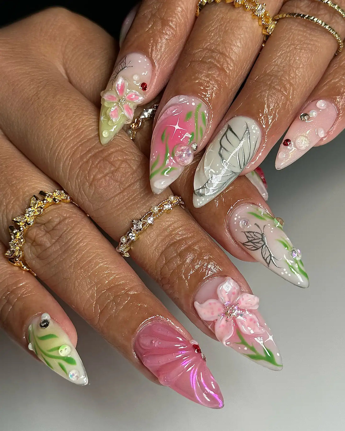 Hand with long stiletto press-on nails featuring pink, green, and white 3D floral and butterfly nail art.