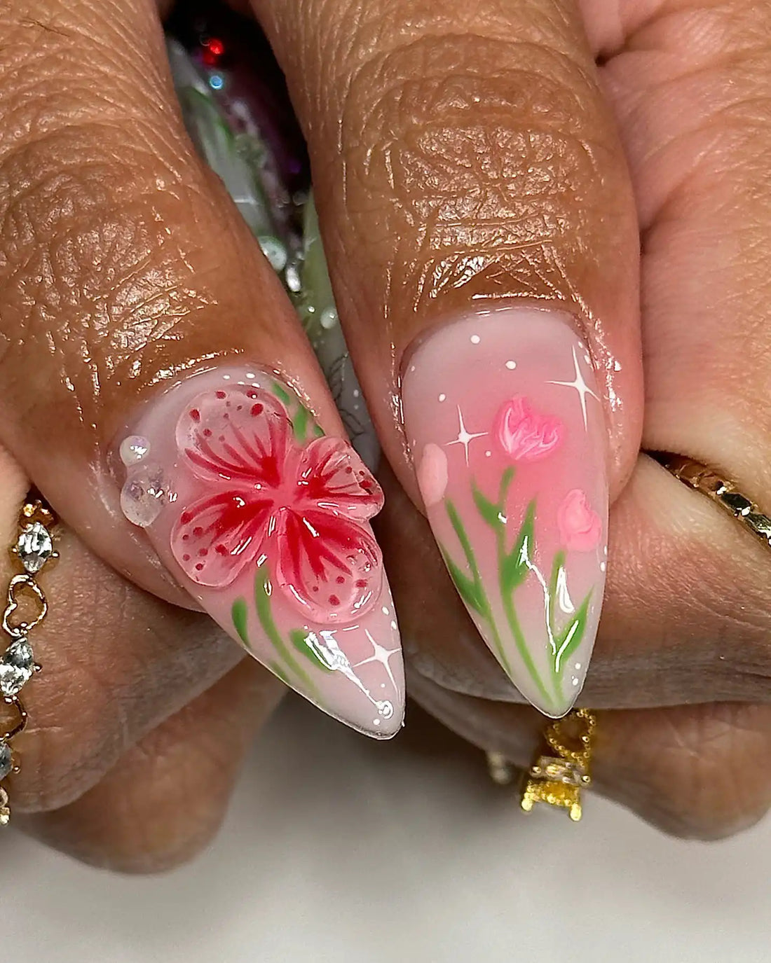 Hand with long stiletto press-on nails featuring pink, green, and white 3D floral and butterfly nail art.