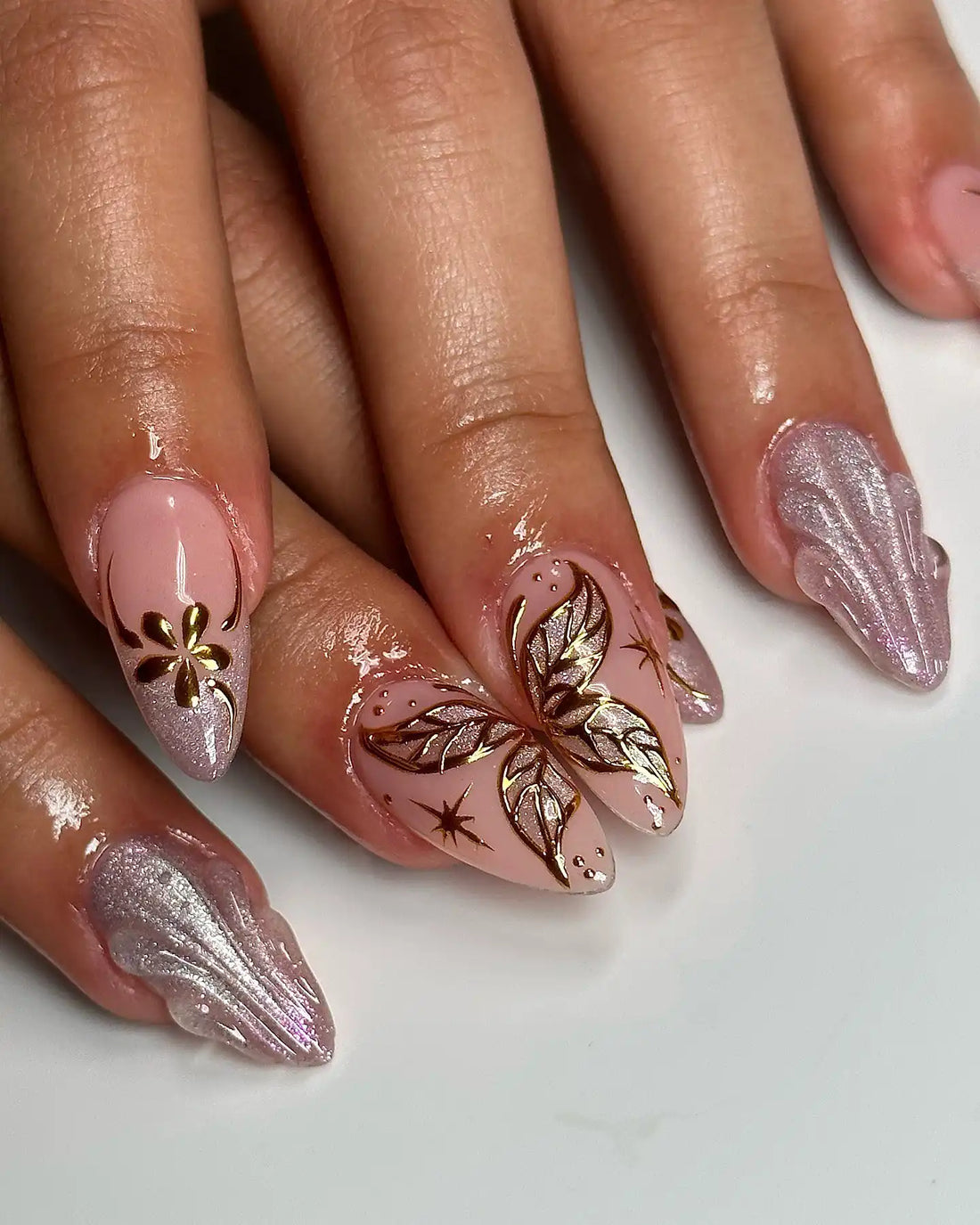 Hand with long pink nude press-on nails featuring gold butterfly and floral designs, chrome shimmer accents.