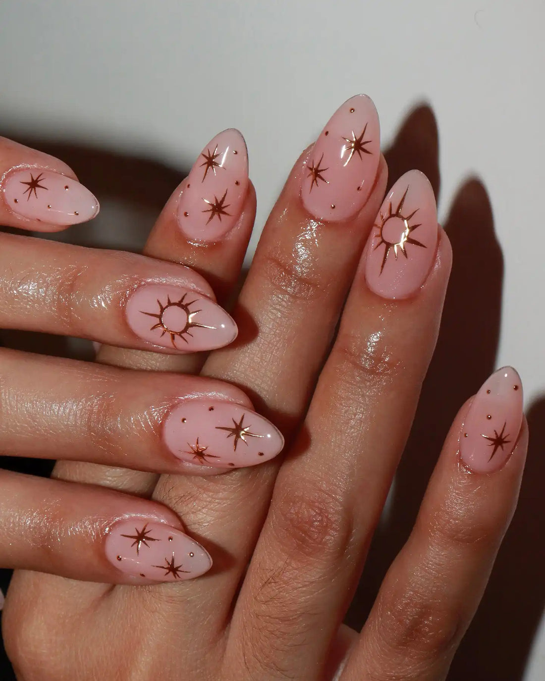 Hand with almond-shaped pink press-on nails featuring gold starburst and constellation designs.