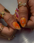 Hand with orange gradient press-on nails, pearl accents, starfish charm, and gold rings