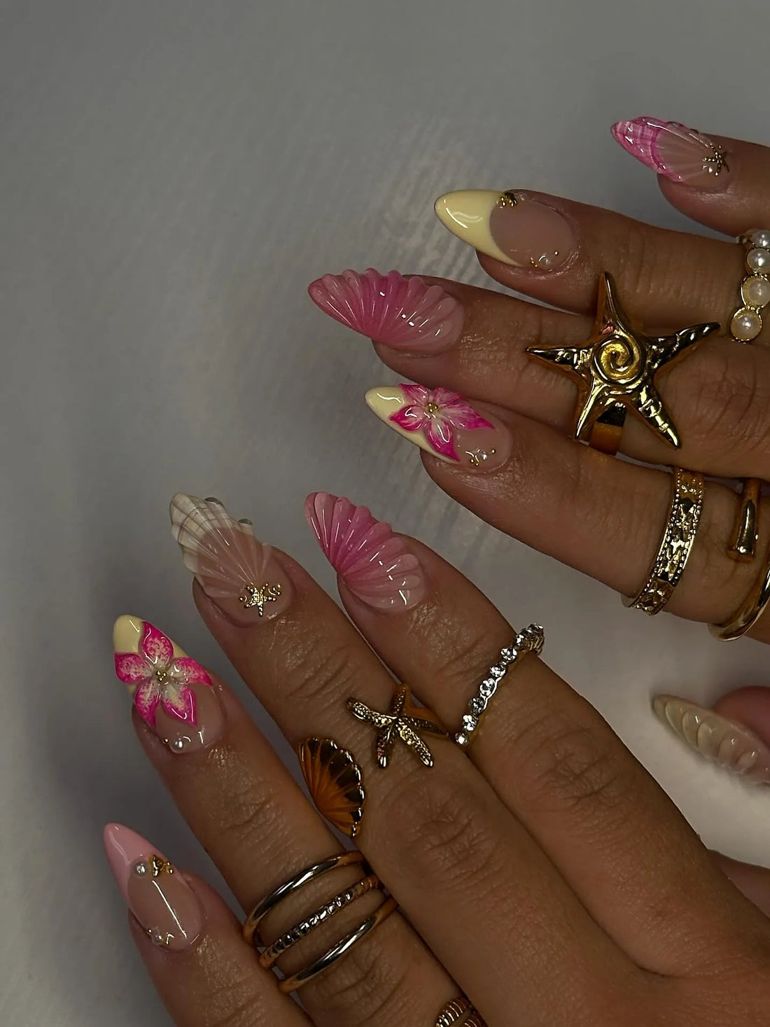 Hand with long press-on nails featuring pink and yellow 3D flowers, shell designs, and gold accents.