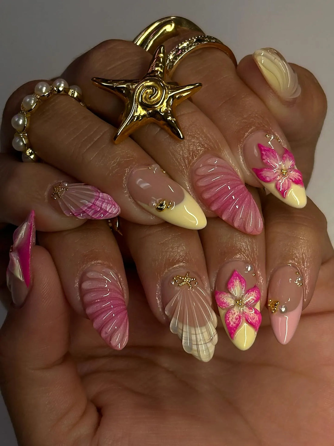 Hand with long press-on nails featuring pink and yellow 3D flowers, shell designs, and gold accents.
