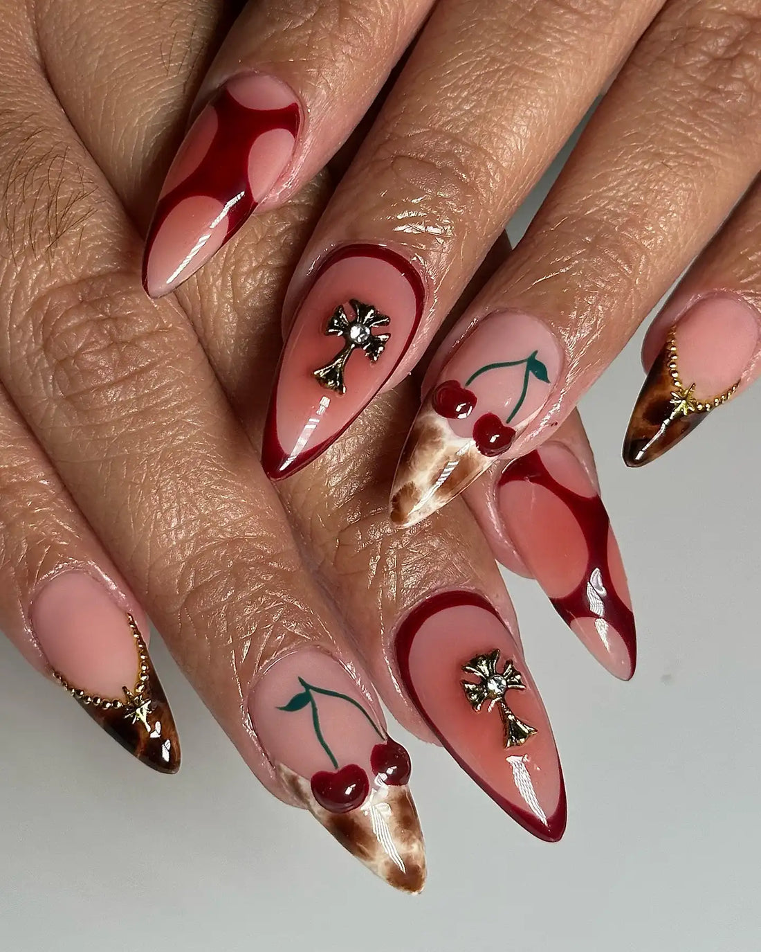 Hand with handmade stiletto press-on nails featuring red cherry, leopard print, cross charms, and chrome accents.