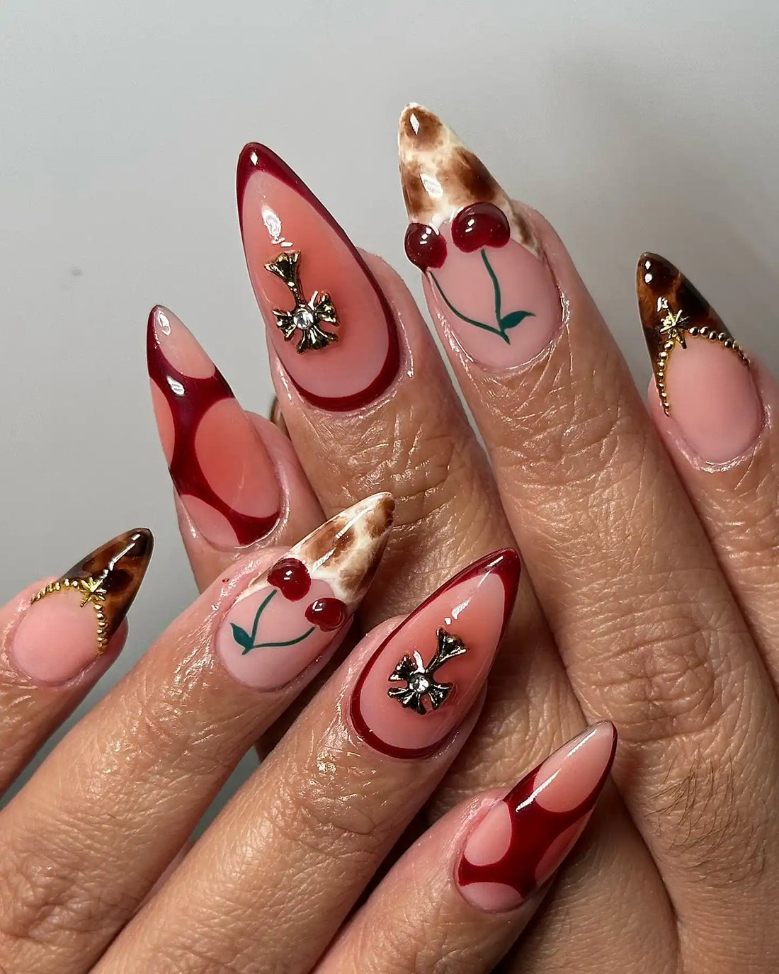 Hand with handmade stiletto press-on nails featuring red cherry, leopard print, cross charms, and chrome accents.