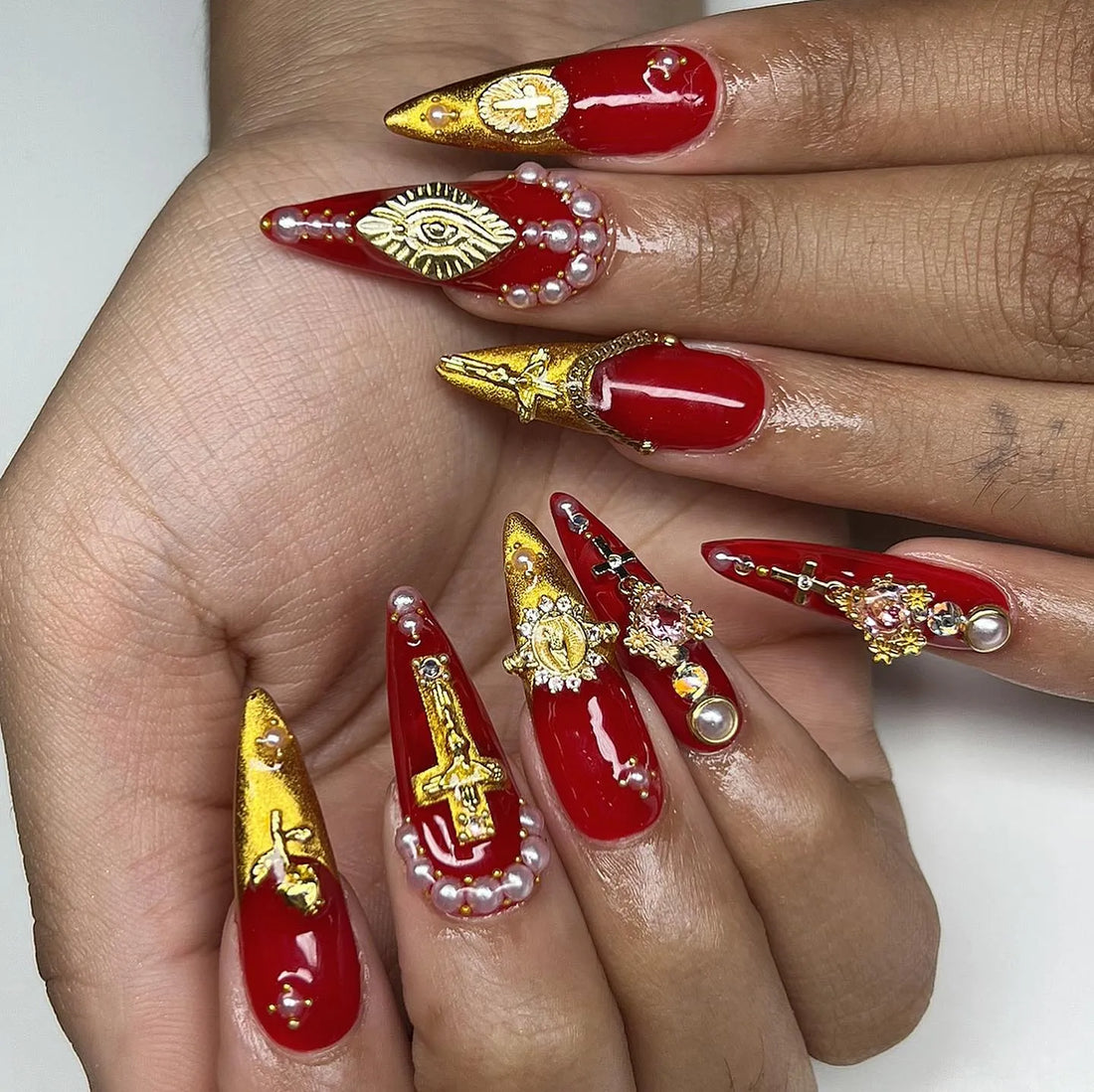 Hand displaying long red press-on nails with gold crosses, evil eye, pearls, and gothic accents.