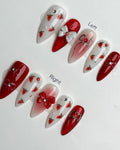 Red and white press-on nails with strawberry fruit art, bow accents, and rhinestone jewels.