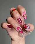 Hand wearing shimmering red and burgundy press-on nails with gemstone and gold embellishments