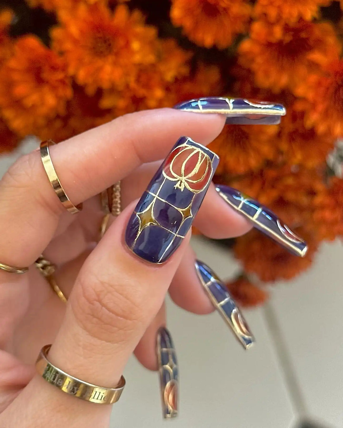 Hand with long press-on nails featuring stained glass pumpkin nail art in gold and orange, autumn flowers background.