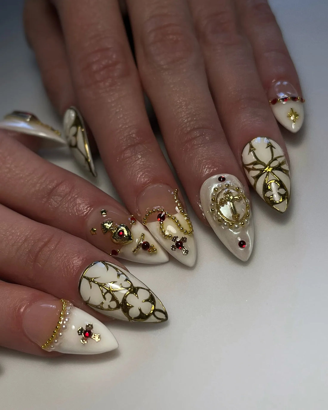 Handmade white stiletto press-on nails with gold gothic cross, pearl, and red gem accents