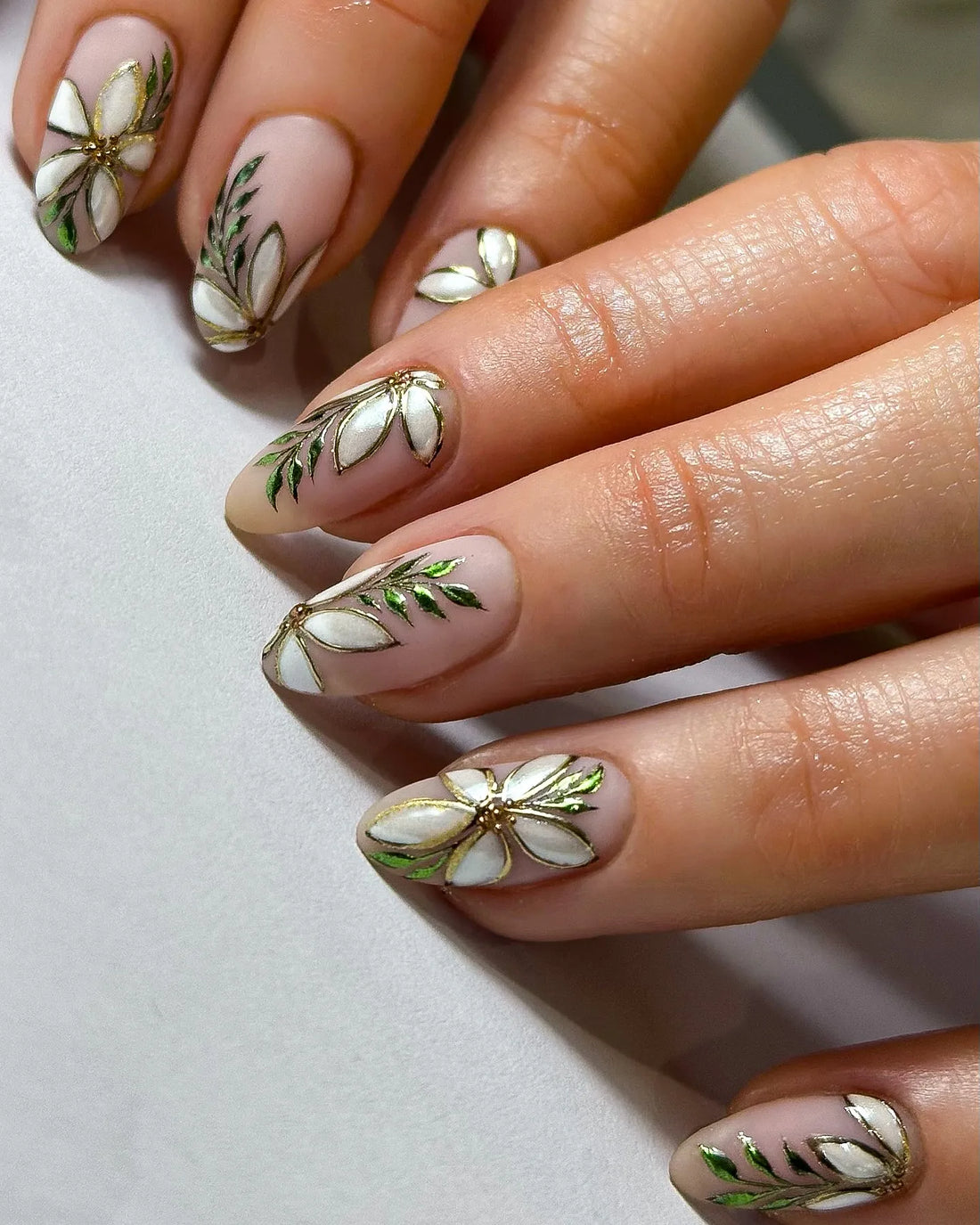 Hand with almond-shaped press-on nails featuring white pearl floral designs and metallic green leaves.