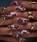 Hand with long white press-on nails featuring pink floral gothic designs, gold accents, and artistic dots