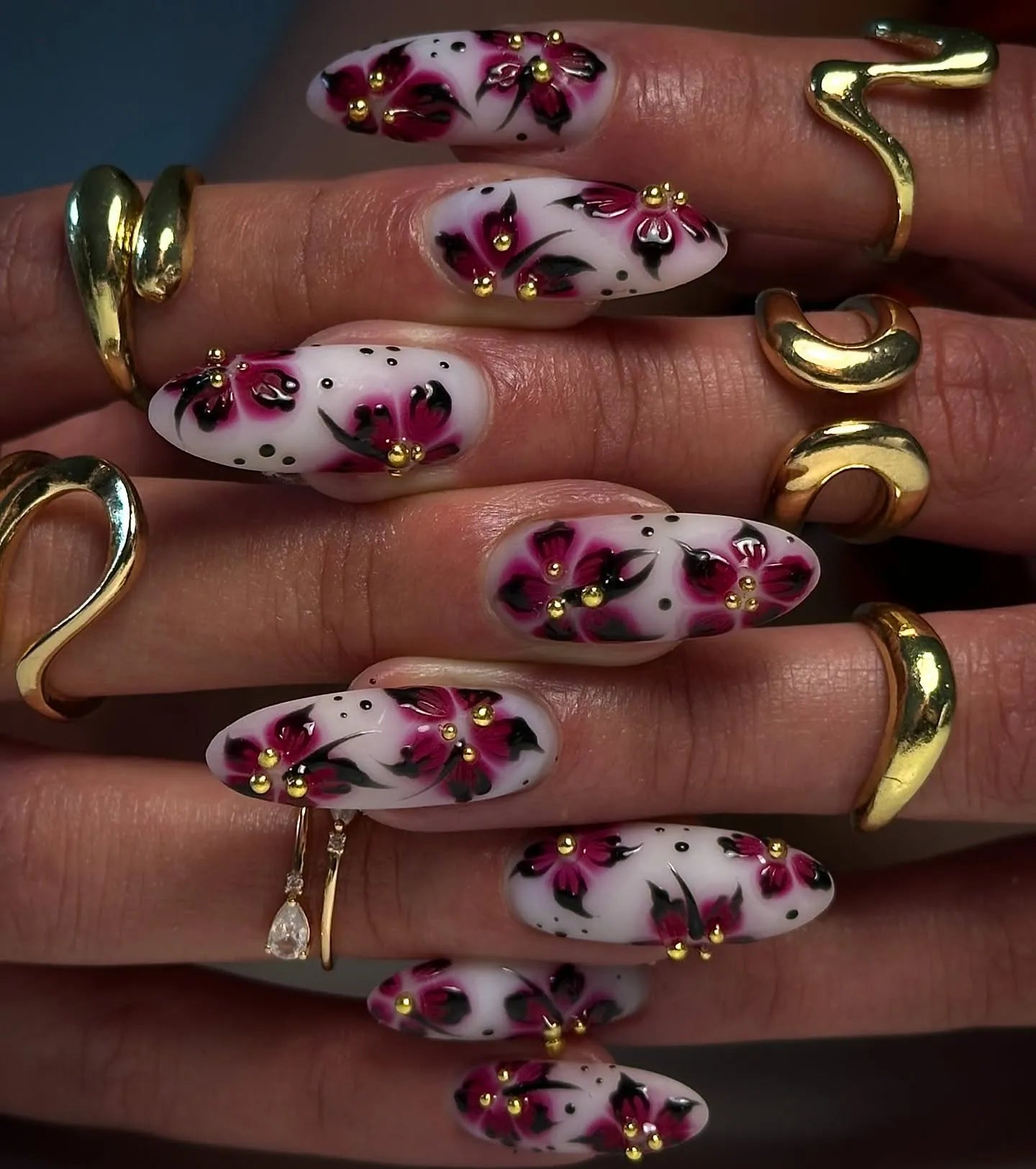 Hand with long white press-on nails featuring pink floral gothic designs, gold accents, and artistic dots