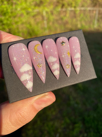Pink press-on nails with white clouds, gold moon and stars, and glitter celestial design