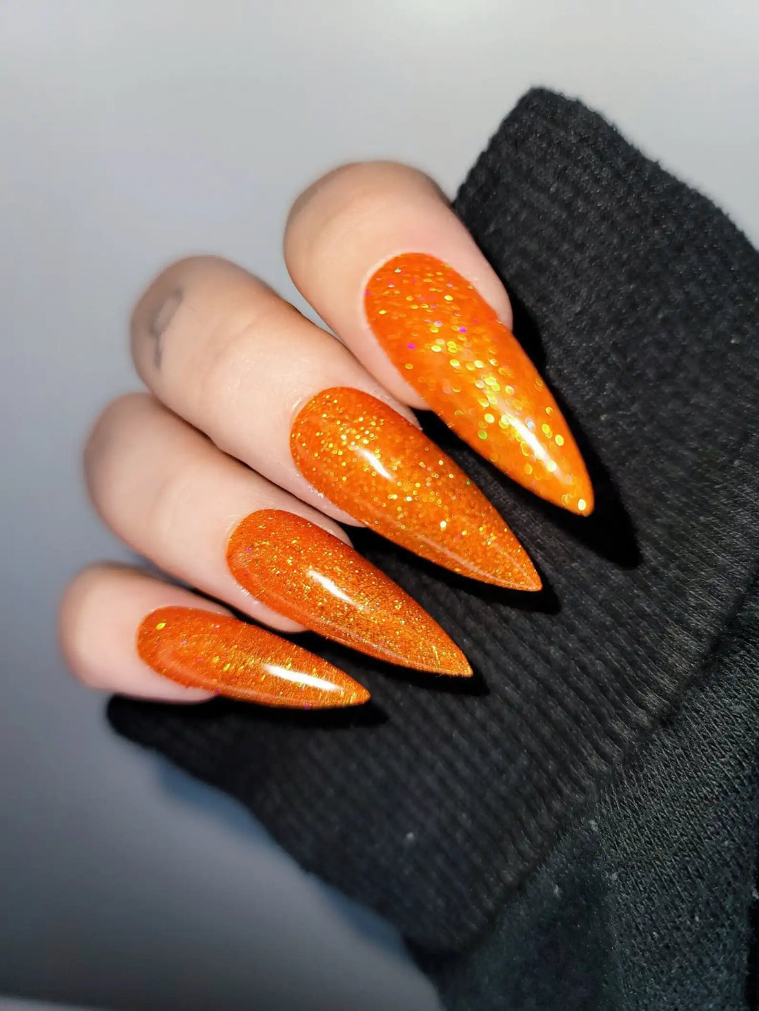 Hand with long stiletto-shaped orange glitter press-on nails, displayed on black sleeve.