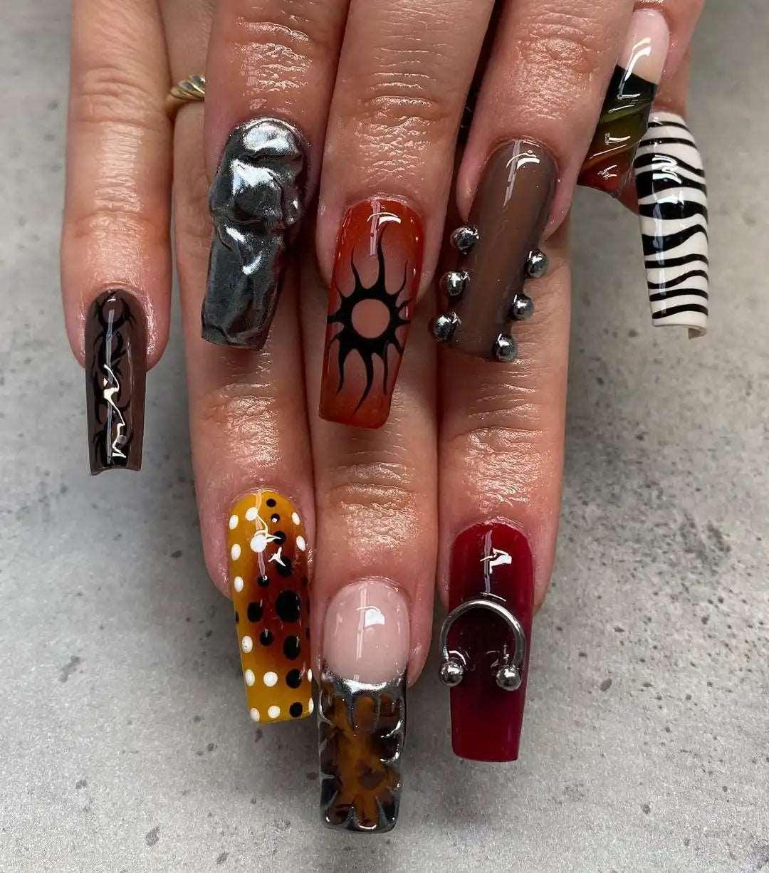 Hand with long press-on nails featuring bold, mixed designs, 3D art, studs, zebra print, and floral patterns.