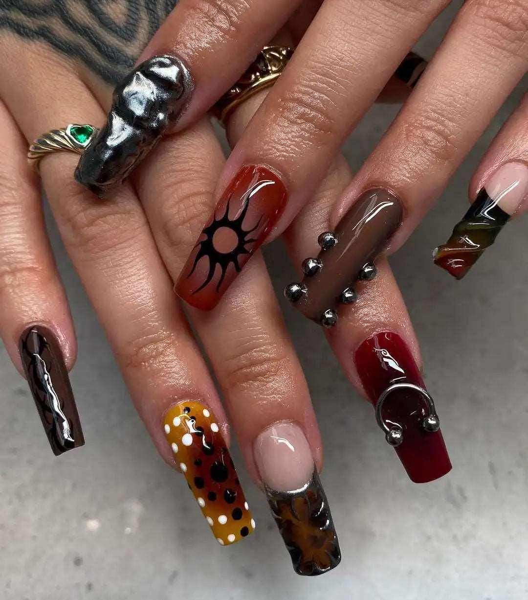 Hand with long press-on nails featuring bold, mixed designs, 3D art, studs, zebra print, and floral patterns.