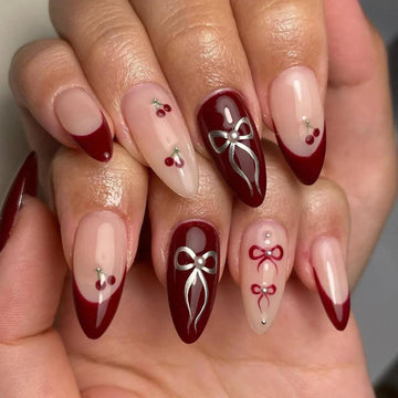 Hand with almond-shaped press-on nails featuring red French tips, cherry and bow designs.