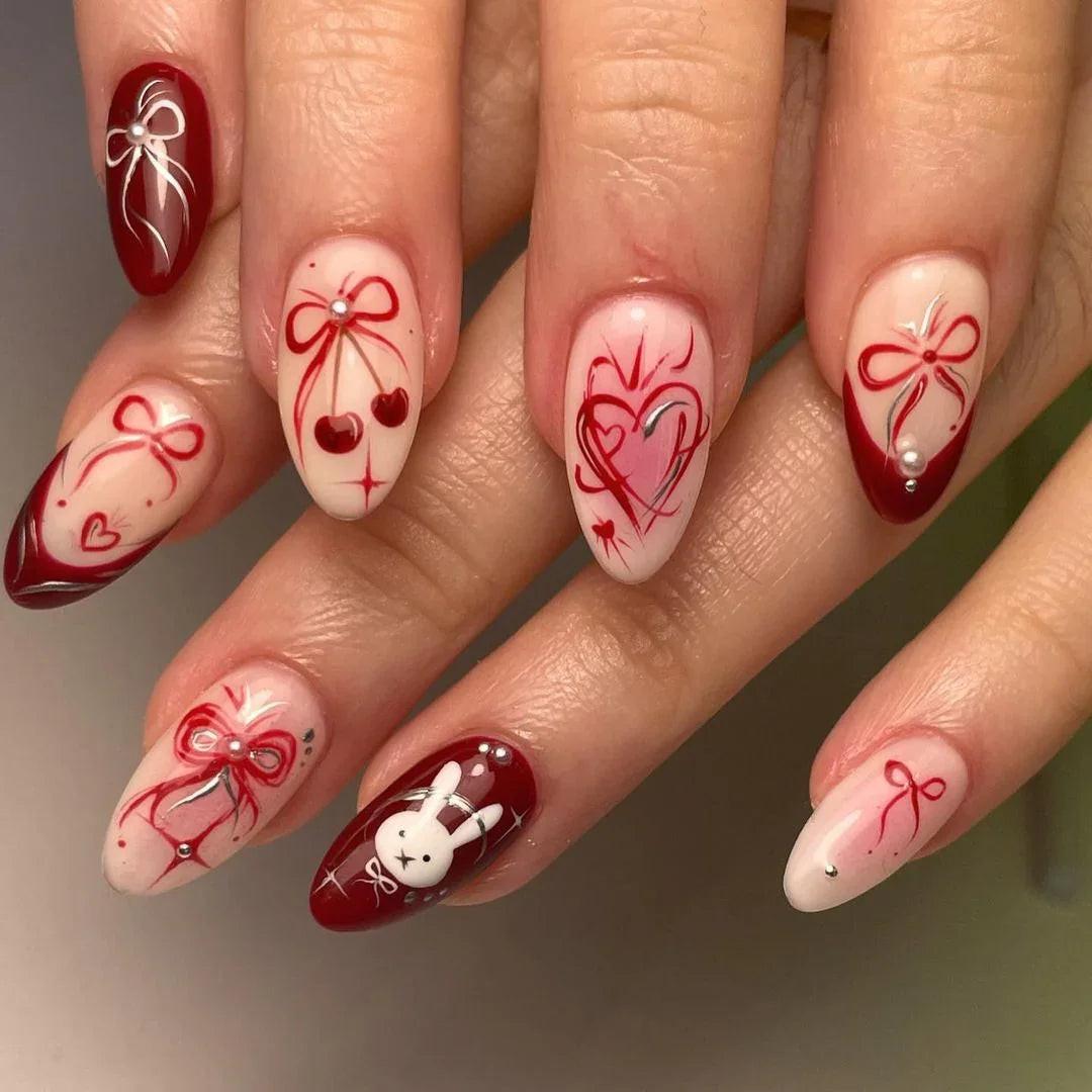 Hand with almond-shaped press-on nails in red and nude tones, featuring heart, bow, cherry, and bunny nail art designs.