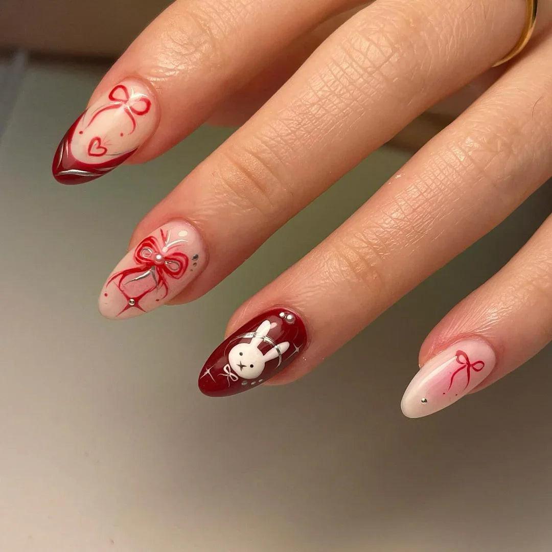 Hand with almond-shaped press-on nails in red and nude, featuring bow and bunny nail art designs.