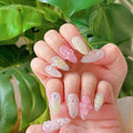 Hand with pastel 3D floral press-on nails in pink and yellow, held in front of green leaves
