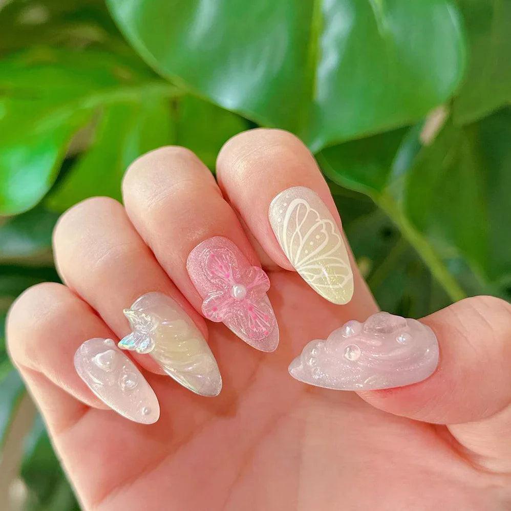 Hand with pastel 3D press-on nails featuring butterfly and flower designs, against green leaves.